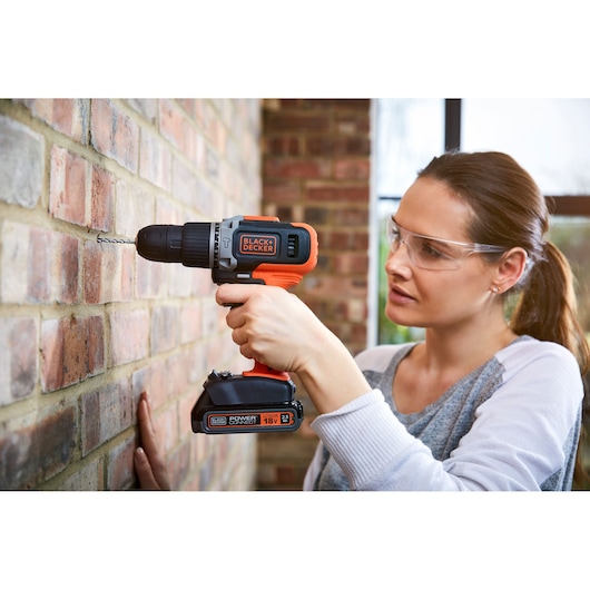 A person using a BLACK+DECKER cordless drill to drill into a brick wall. The individual’s face is blurred for privacy. The drill is orange and black, and the scene is set indoors.