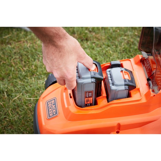 BLACK+DECKER BCMW33184L2 lawn mower close-up of hand inserting 18V battery into orange battery compartment.