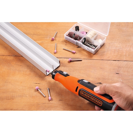 A person uses a BLACK+DECKER rotary tool to work on a metal piece, with various rotary tool accessories and attachments placed on a wooden surface.