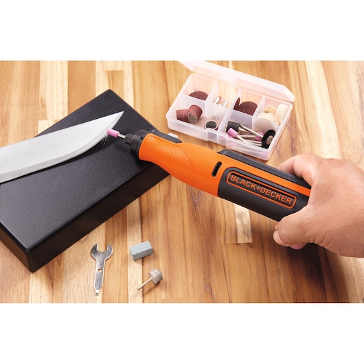 A hand holding a BLACK+DECKER rotary tool is sharpening a large knife blade on a black block. Several tool accessories and attachments are visible, including a wrench, various grinding and sanding bits, and a storage box, all set on a wooden surface.