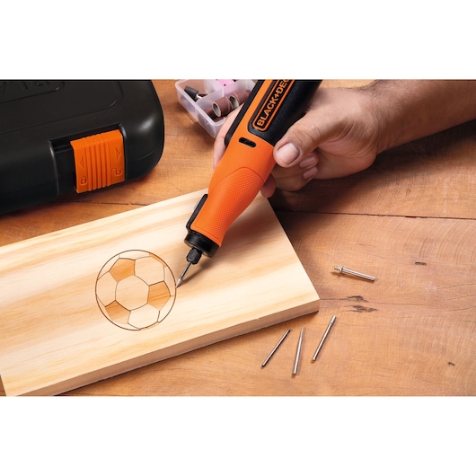 A BLACK+DECKER rotary tool is being used to engrave the outline of a soccer ball onto a piece of wood. Nearby are some rotary tool accessories and a storage case.