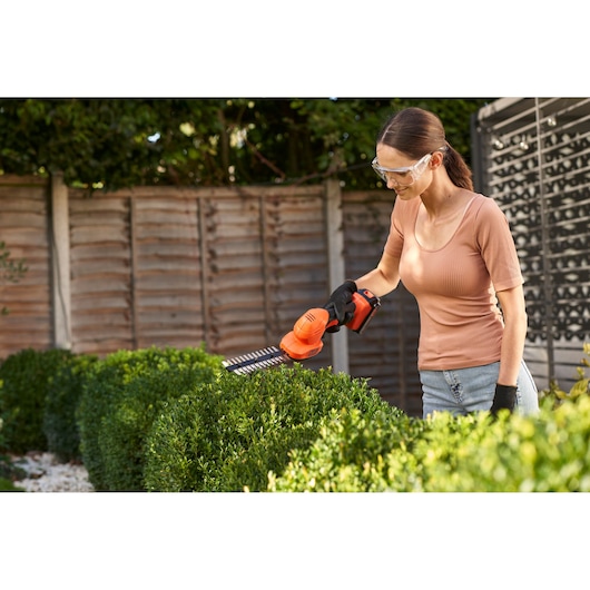A person is using an orange BLACK+DECKER cordless hedge trimmer, model BCSS18B, to trim green hedges in a garden. The user is wearing gloves and protective eyewear.