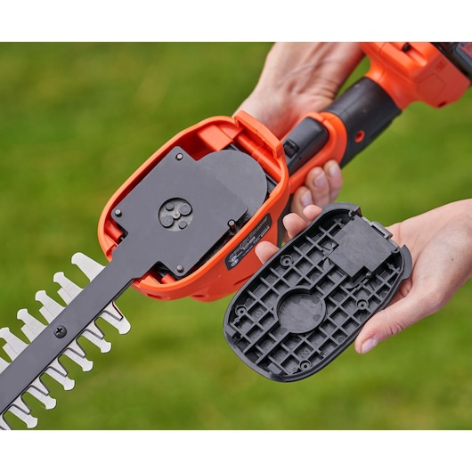 Close-up of a BLACK+DECKER cordless shrub and grass shear with the casing removed, showing the internal mechanism and blade attachment. A person holds the detached casing cover in their hand.