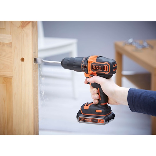 A person using a BLACK+DECKER 18V cordless drill to make a hole in a wooden door.