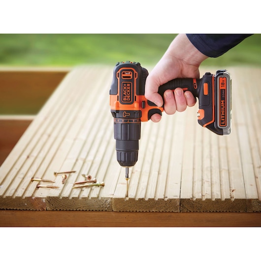 A hand holding a BLACK+DECKER cordless lithium 18V drill, screwing into a wooden surface with several screws lying nearby.