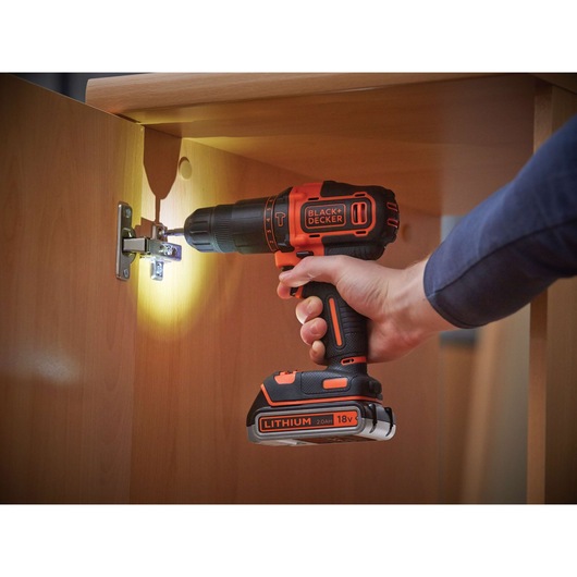 A person using a BLACK+DECKER 18V cordless drill with built-in light to install a hinge inside a wooden cabinet.