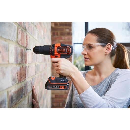 A person using a BLACK+DECKER 18V cordless drill to drill into a brick wall. The drill is held firmly, and the person's face is blurred for privacy.