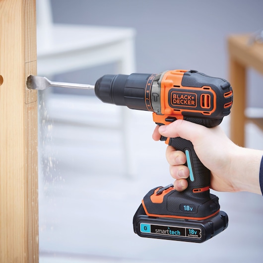 A hand holding a BLACK+DECKER cordless drill with an 18V lithium battery, drilling a hole into a wooden surface.