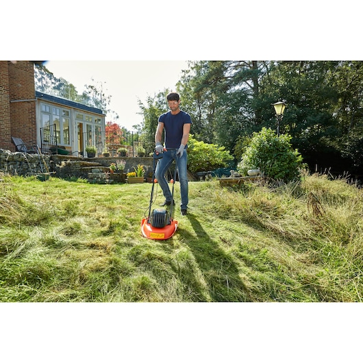 A person mowing an overgrown grassy lawn with an orange BLACK+DECKER electric lawn mower in a garden next to a house.