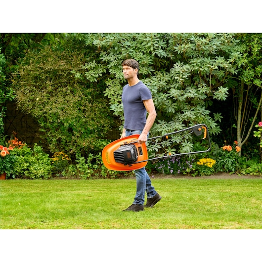 A person carrying an orange BLACK+DECKER lawn mower across a grassy garden area, with flowers and large green shrubs in the background.