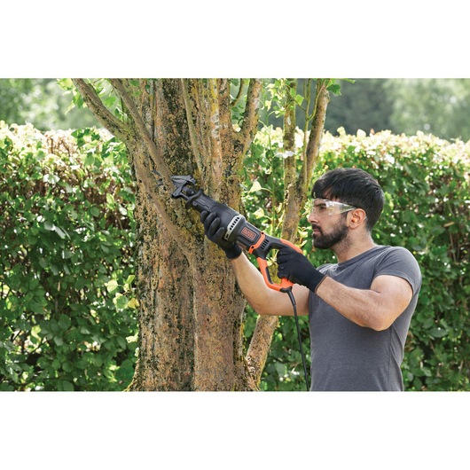 Person using BLACKDECKER BES301K reciprocating saw to cut tree branch in outdoor garden.