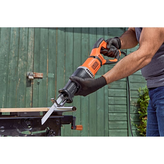 A person wearing gloves is using a BLACK+DECKER BES301 reciprocating saw to cut through a metal pipe, with the saw positioned on a workbench outdoors.