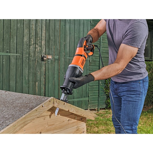A person using a BLACK+DECKER BES301 reciprocating saw to cut a piece of wood outdoors, wearing gloves and holding the saw on a wooden structure.