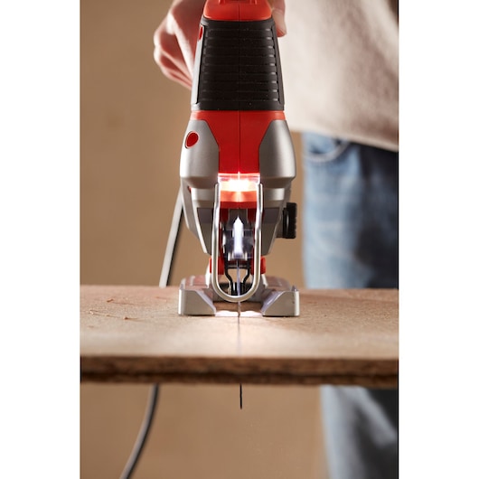 Close-up view of a BLACK+DECKER electric jigsaw cutting through a wooden plank, held by a person.
