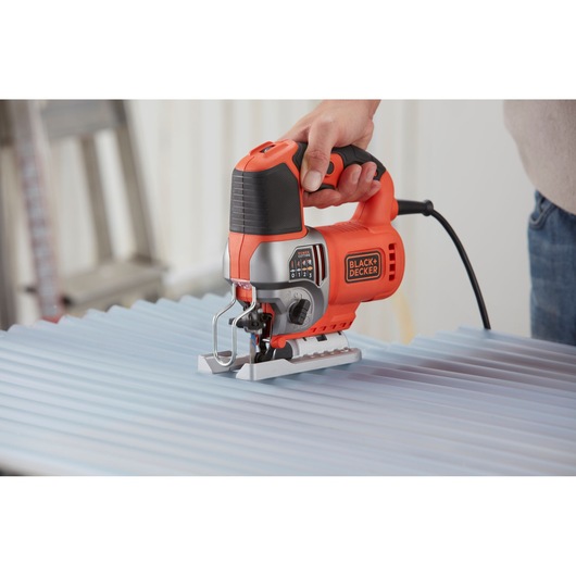 A person using a BLACK+DECKER BES610 electric jigsaw to cut a corrugated metal sheet. The jigsaw is orange and black, held firmly by hand, and plugged in for use.