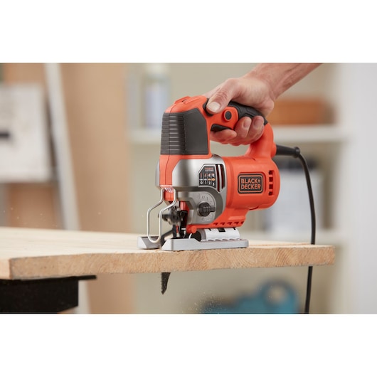 A close-up view of a BLACK+DECKER electric jigsaw being used to cut a piece of wood. The tool is held by a person's hand and is plugged into power.