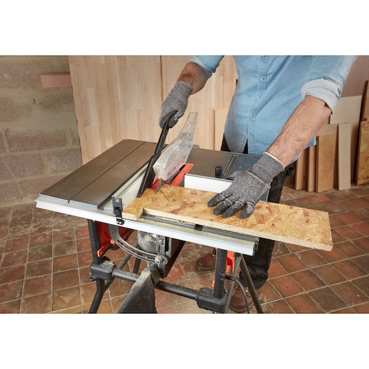 A person wearing protective gloves is using a BLACK+DECKER BES720 table saw to cut a piece of oriented strand board in a workshop.