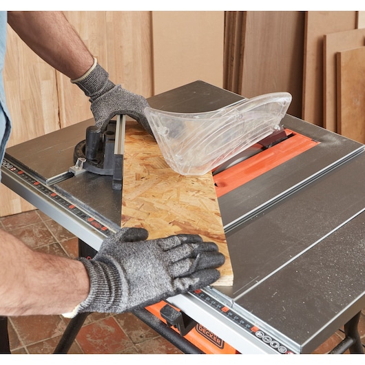 A person wearing protective gloves operates a BLACK+DECKER table saw to cut a piece of plywood, with a transparent blade guard for safety.