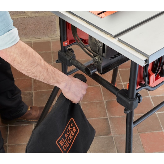 A person is attaching a BLACK+DECKER branded dust collection bag to a table saw. The table saw is mounted on a stand, with visible wiring and components, placed on a tiled floor.