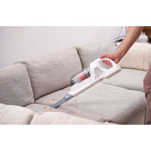 A person uses a white handheld vacuum cleaner to clean crumbs from a light grey sofa.