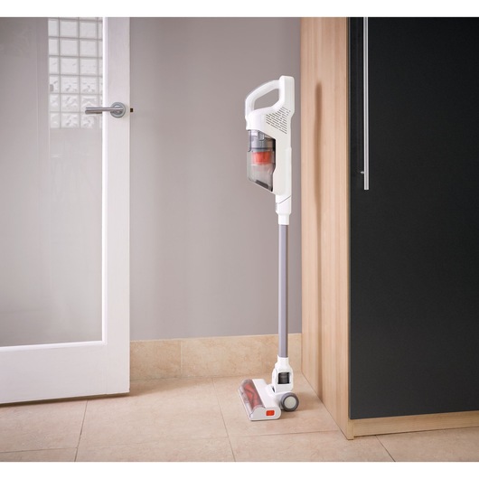 A white cordless stick vacuum cleaner with a transparent dust container is standing upright against a wall in a modern kitchen setting.
