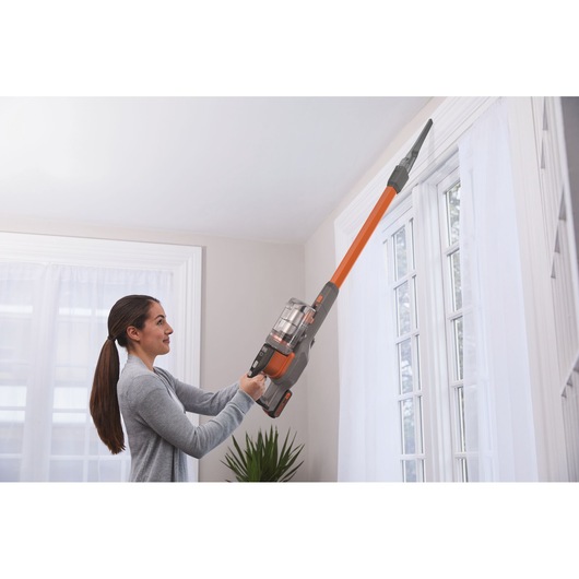 A person uses a BLACK+DECKER cordless stick vacuum cleaner with an extended nozzle to clean the upper edge of a window inside a bright room. The vacuum model BHFEV182C is being operated vertically.