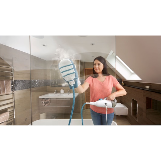 A person using a steam cleaning glove attached to a BLACK+DECKER steam cleaner to clean a glass shower door in a modern bathroom. The person's face is blurred for privacy.