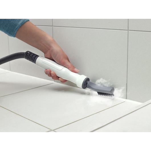 A hand is using a steam cleaning tool with a brush attachment to clean the grout lines between white tiles.