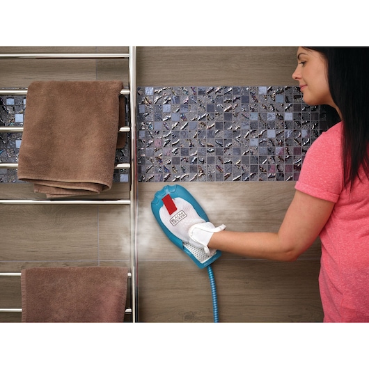 A person using a BLACK+DECKER steam cleaning glove to clean dark mosaic tiles on a bathroom wall. Brown towels are hanging on a metal towel rack nearby.