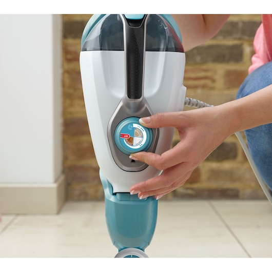 Close-up view of a person adjusting the dial on a BLACK+DECKER steam mop, with the device positioned upright on a tiled floor.