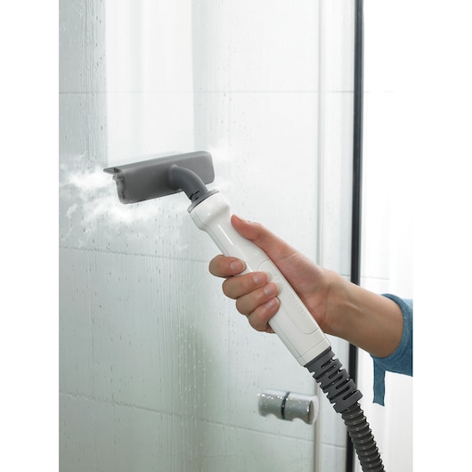 A person is using a BLACK+DECKER steam cleaner to clean a glass shower door. The device is releasing steam onto the glass surface.
