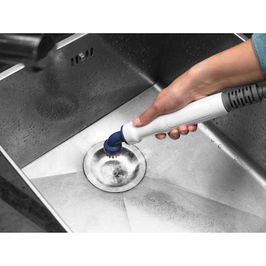 A hand holding a BLACK+DECKER cleaning tool with a brush attachment, scrubbing the drain area of a stainless steel sink.