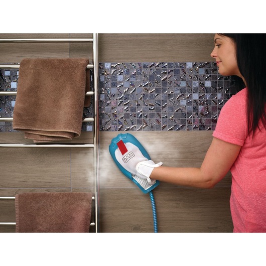 A person using a BLACK+DECKER steam cleaning glove appliance to clean a tiled wall in a bathroom. The steam cleaner emits vapor, and two brown towels hang on a heated towel rack nearby. The person's face is not visible.