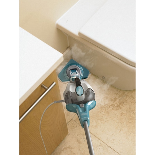 A BLACK+DECKER steam mop cleaning the floor in a bathroom, with steam visible as it cleans the area between a cabinet and a toilet.
