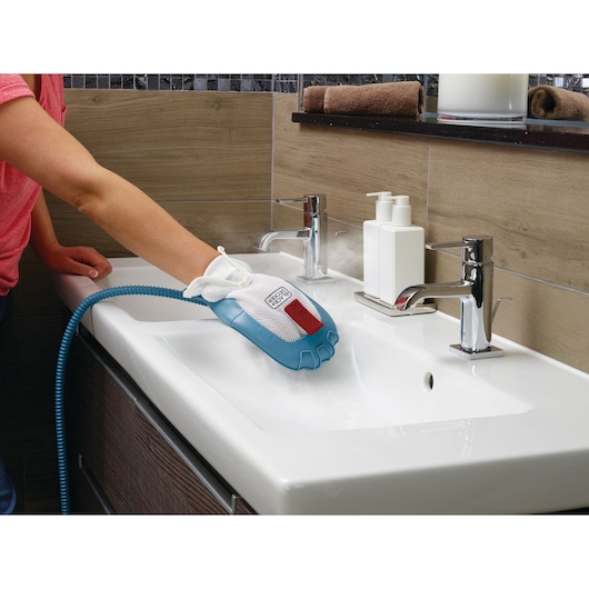 A person is using a BLACK+DECKER steam cleaning glove to clean a white bathroom sink, with steam visible around the glove. The bathroom has modern fixtures and two soap dispensers on the sink.