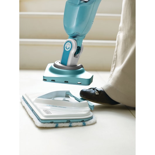 A person uses their foot to attach a cleaning pad to a BLUE BLACK+DECKER steam mop on a light-colored tile floor.