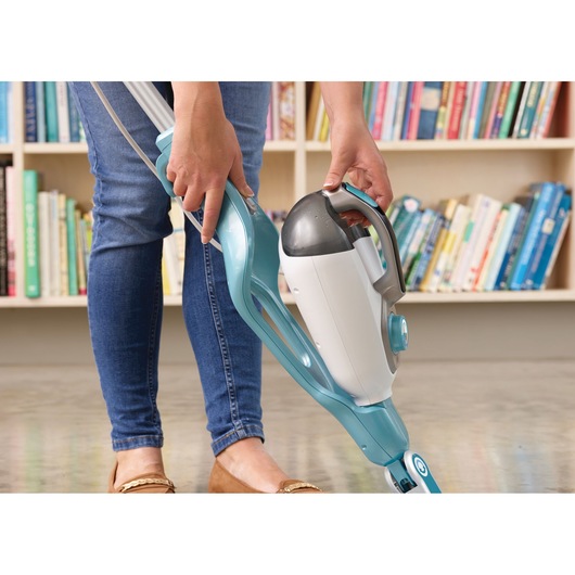 A person is using a blue and white BLACK+DECKER steam mop on a hard floor in a room with a bookshelf filled with colorful books.