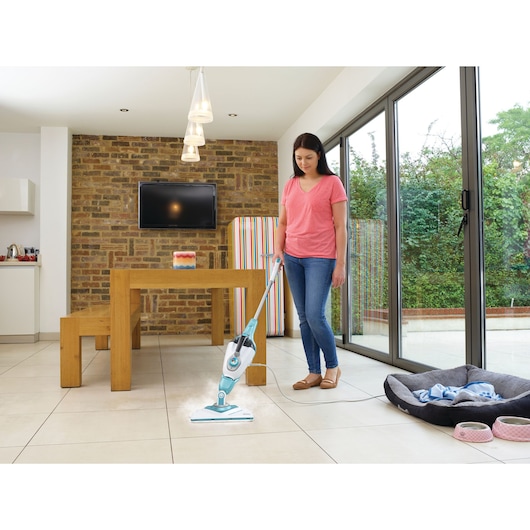 A person using a BLACK+DECKER steam mop to clean tile flooring in a modern living space with a wooden table, benches, and large glass doors leading to a garden.