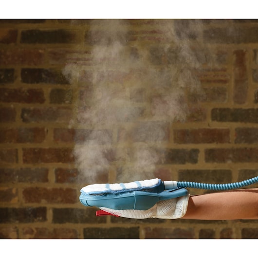 A close-up of a hand wearing a steam cleaning glove, emitting visible steam, in front of a brick wall background.