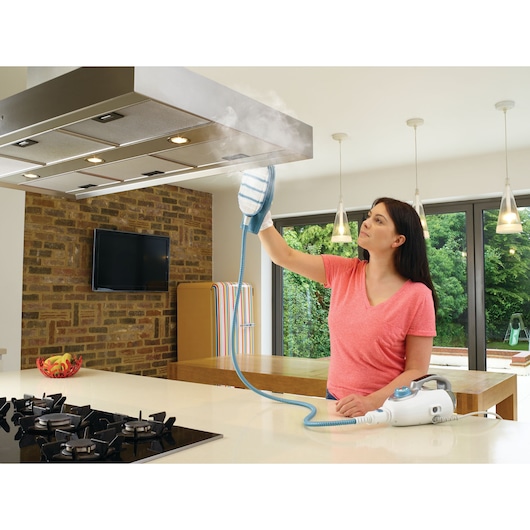 A person using a BLACK+DECKER steam cleaner (model BHSM168DSM) to clean a kitchen range hood. The steam cleaner is white with blue accents, and the kitchen is modern with brick and glass elements.