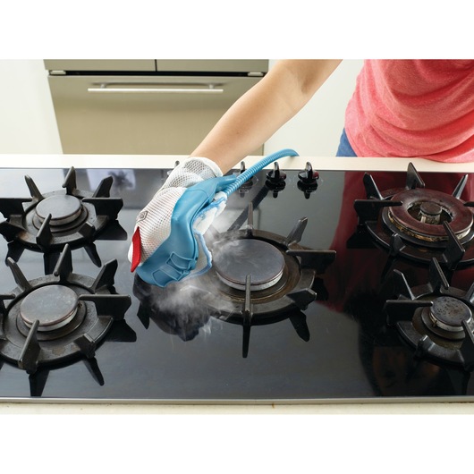 A person uses a steam cleaning glove to clean a black gas stove top. Steam is visible coming from the glove during cleaning.