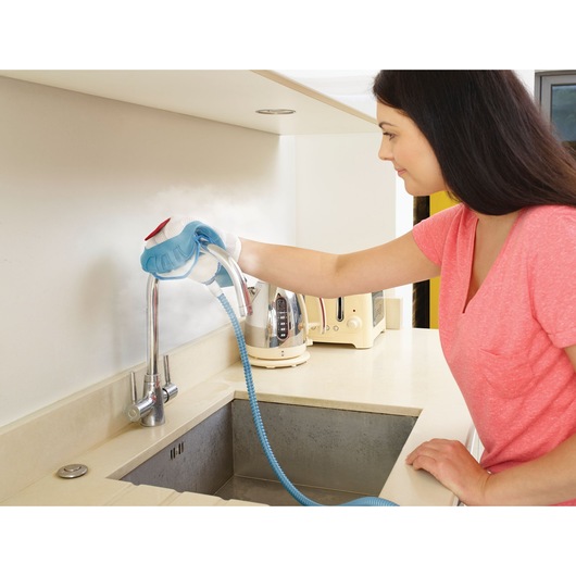 Person using a steam cleaning device to clean a kitchen faucet and backsplash. The device has a blue handle and is producing visible steam.