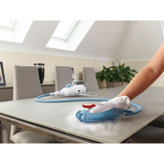 A steam cleaning glove attachment connected to a BLACK+DECKER steam cleaner is being used to clean a tabletop in a modern dining room.