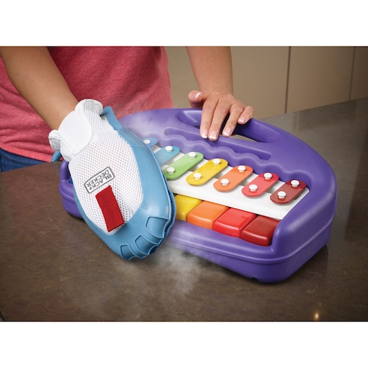 A person uses a BLACK+DECKER steam cleaning glove to clean a colorful children's xylophone toy placed on a countertop.