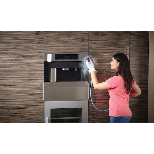 A person is using a BLACK+DECKER steam cleaning glove to clean the exterior of a built-in kitchen appliance. The steam is visibly coming out of the glove while the person wears it and cleans the appliance's front surface.