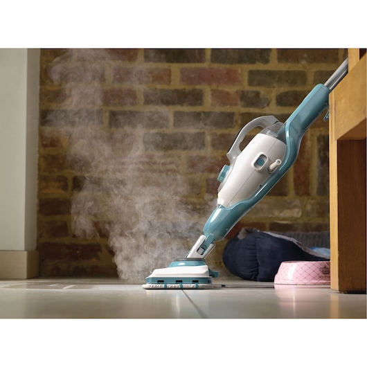 A BLACK+DECKER steam mop cleaning a tiled floor, emitting steam from its cleaning head. The mop is positioned under a piece of furniture with a brick wall in the background.