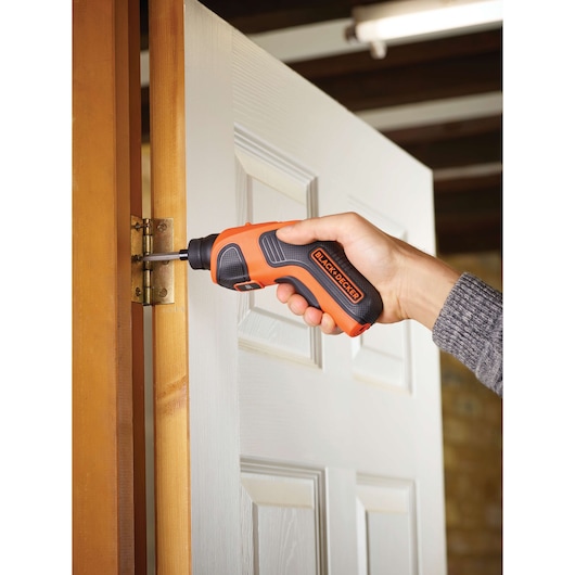 A person uses a BLACK+DECKER cordless screwdriver to fasten a hinge on a wooden door frame.