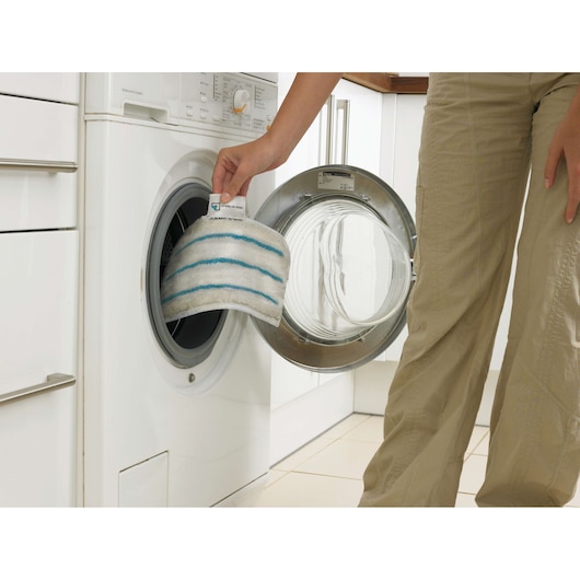 A person is placing a white cleaning pad with blue stripes into the drum of a front-loading washing machine.