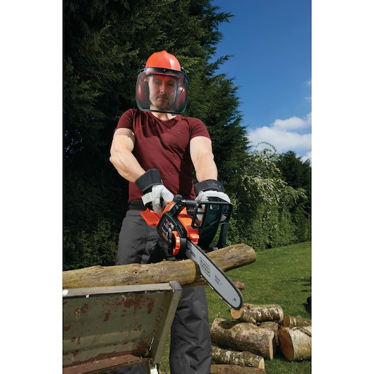 A person wearing protective gear, including a helmet and face shield, is using a BLACK+DECKER cordless chainsaw (model GKC1825L20) to cut logs outdoors. The person is working in a garden with green trees and blue sky in the background.