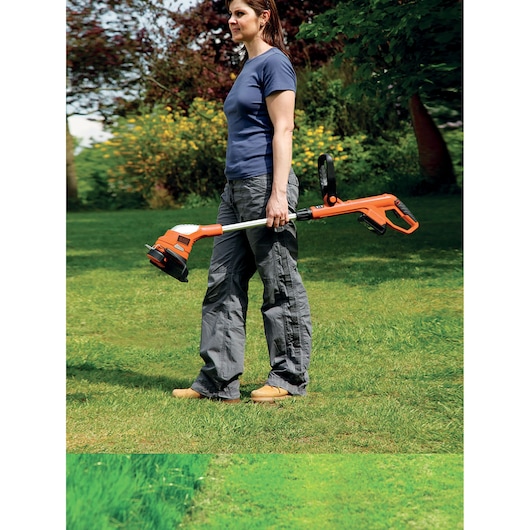 A person standing outdoors on grass, holding an orange and black BLACK+DECKER cordless grass trimmer.